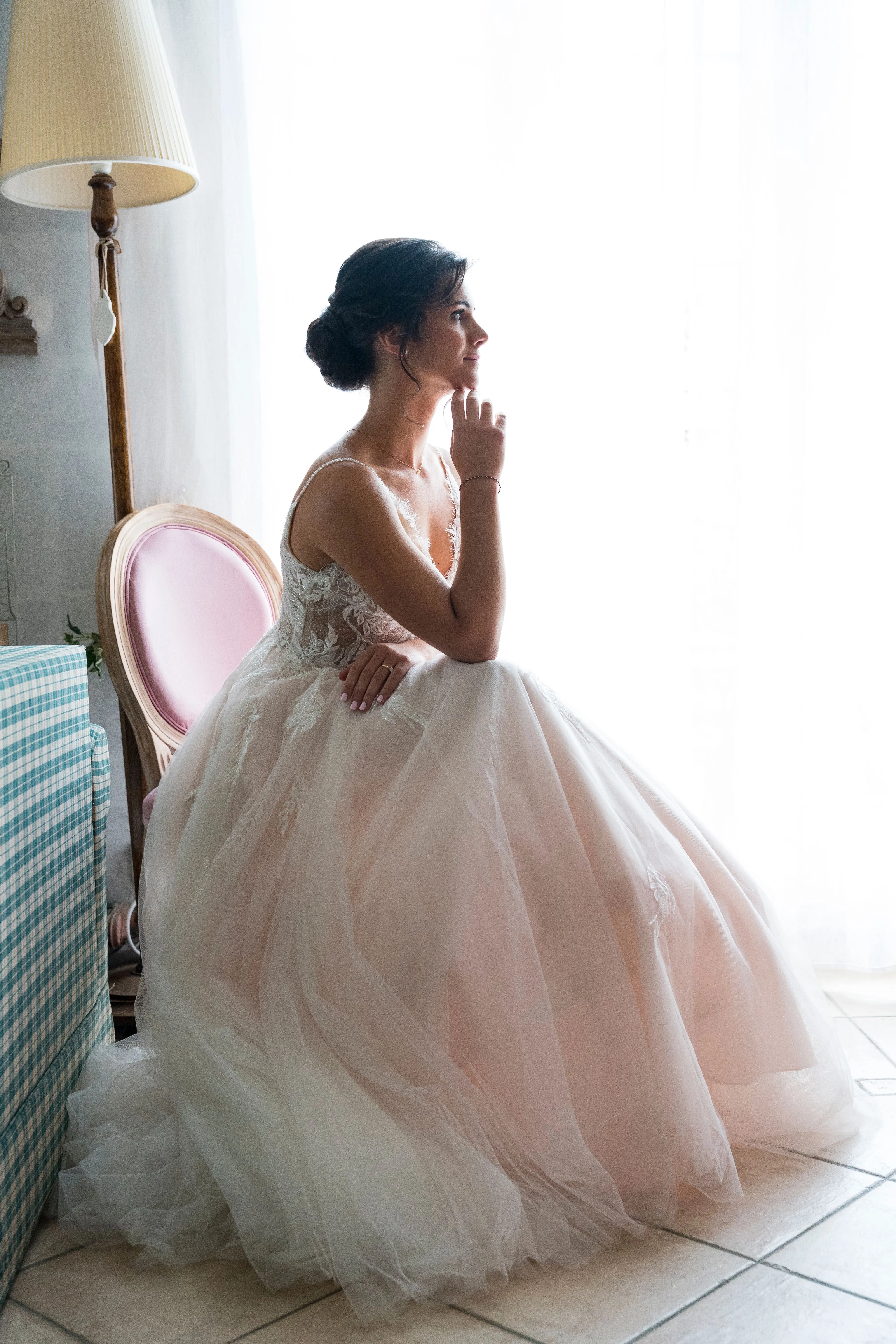 Mariée en robe rose poudré, assise près d'une fenêtre lumineuse