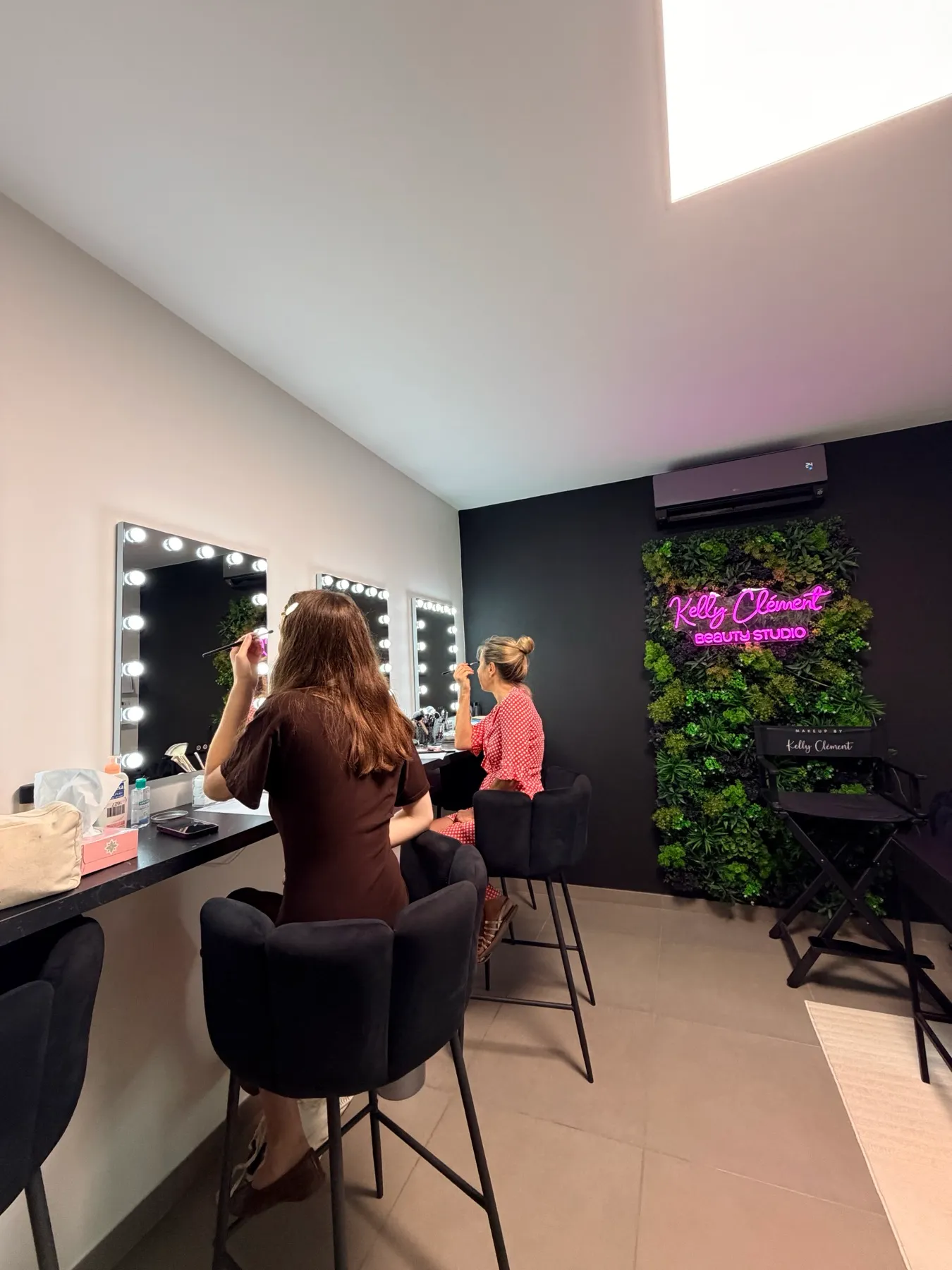 Deux clientes en cours de maquillage à leur poste, devant l'enseigne lumineuse Kelly Clément Beauty Studio